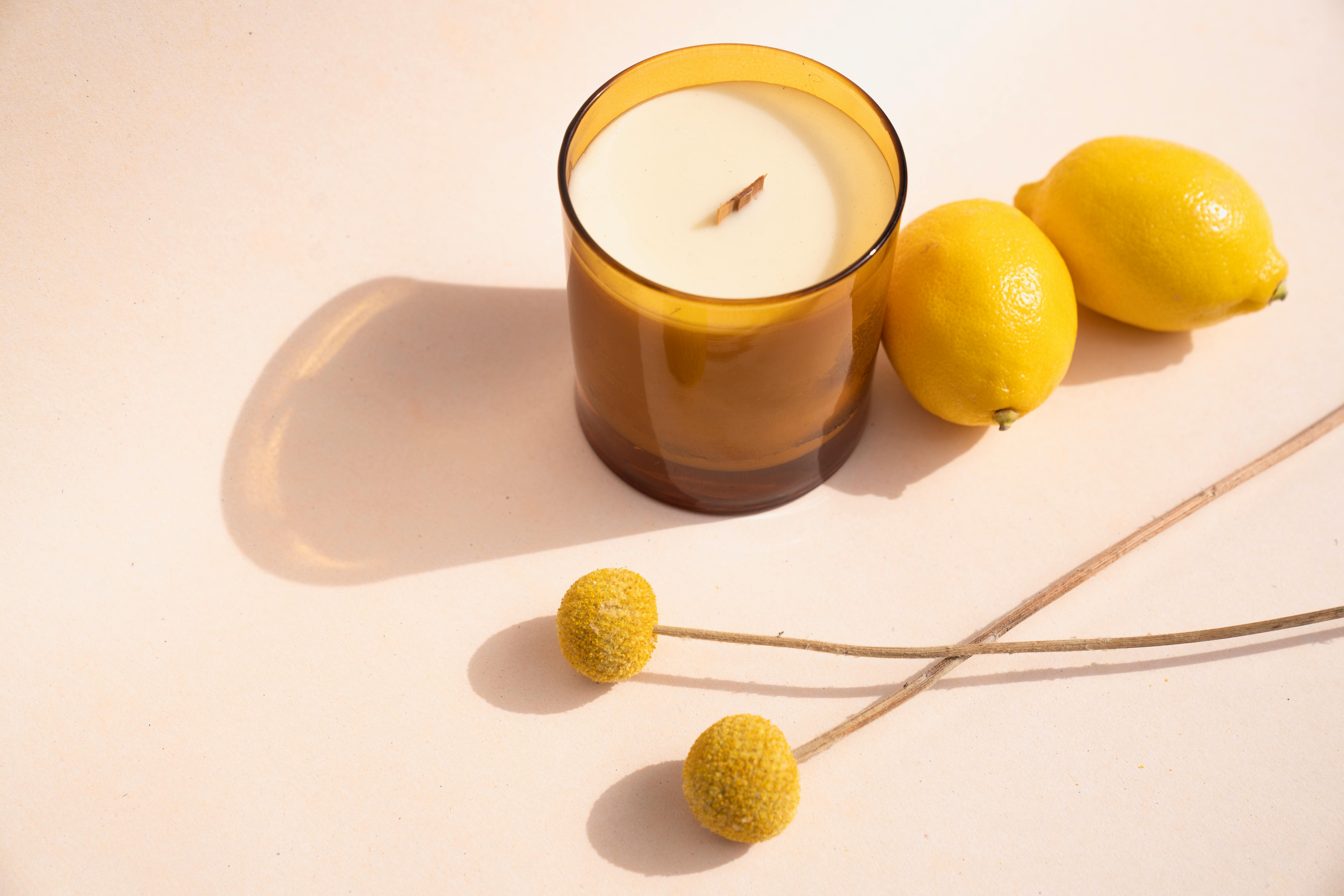 Scented candle in a brown glass holder beside lemons and yellow flowers on a white surface.